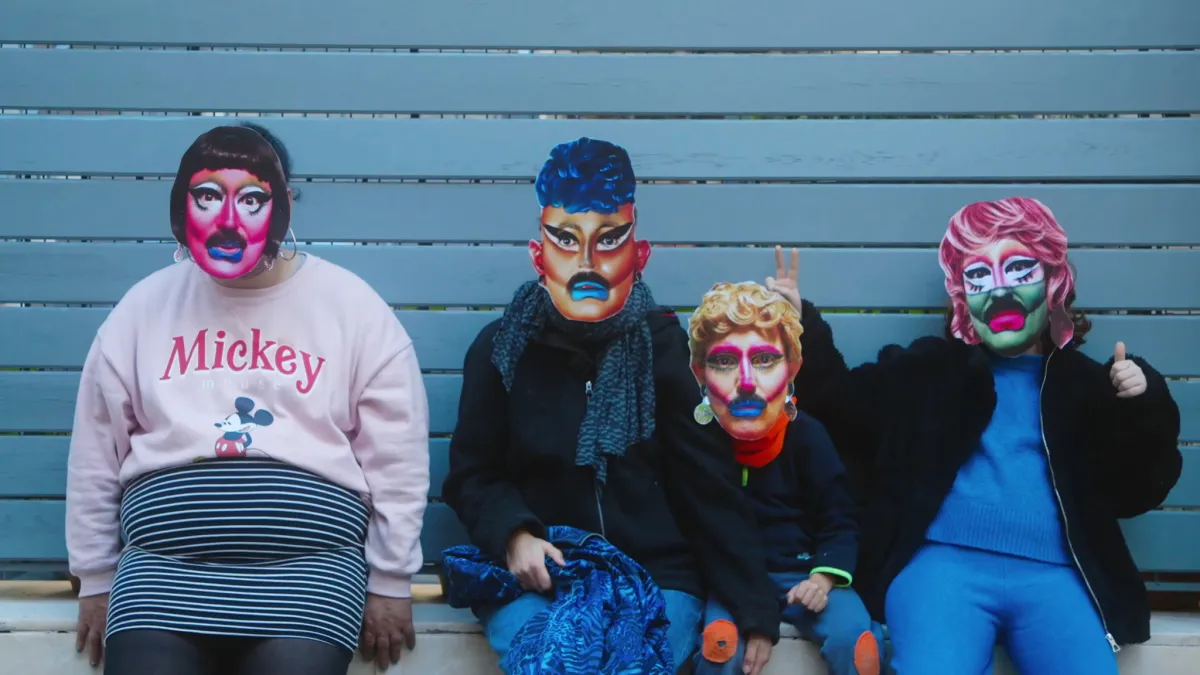 Four people sit against a light blue wall wearing regular clothes and paper masks of drag queens