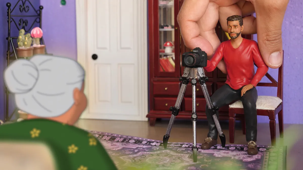 A figurine of a brown man with brown hair and beard in a red shirt and black pants is being placed by a hand in front of a camera on a tripod facing a 2D animated old woman with white hair seen from the back
