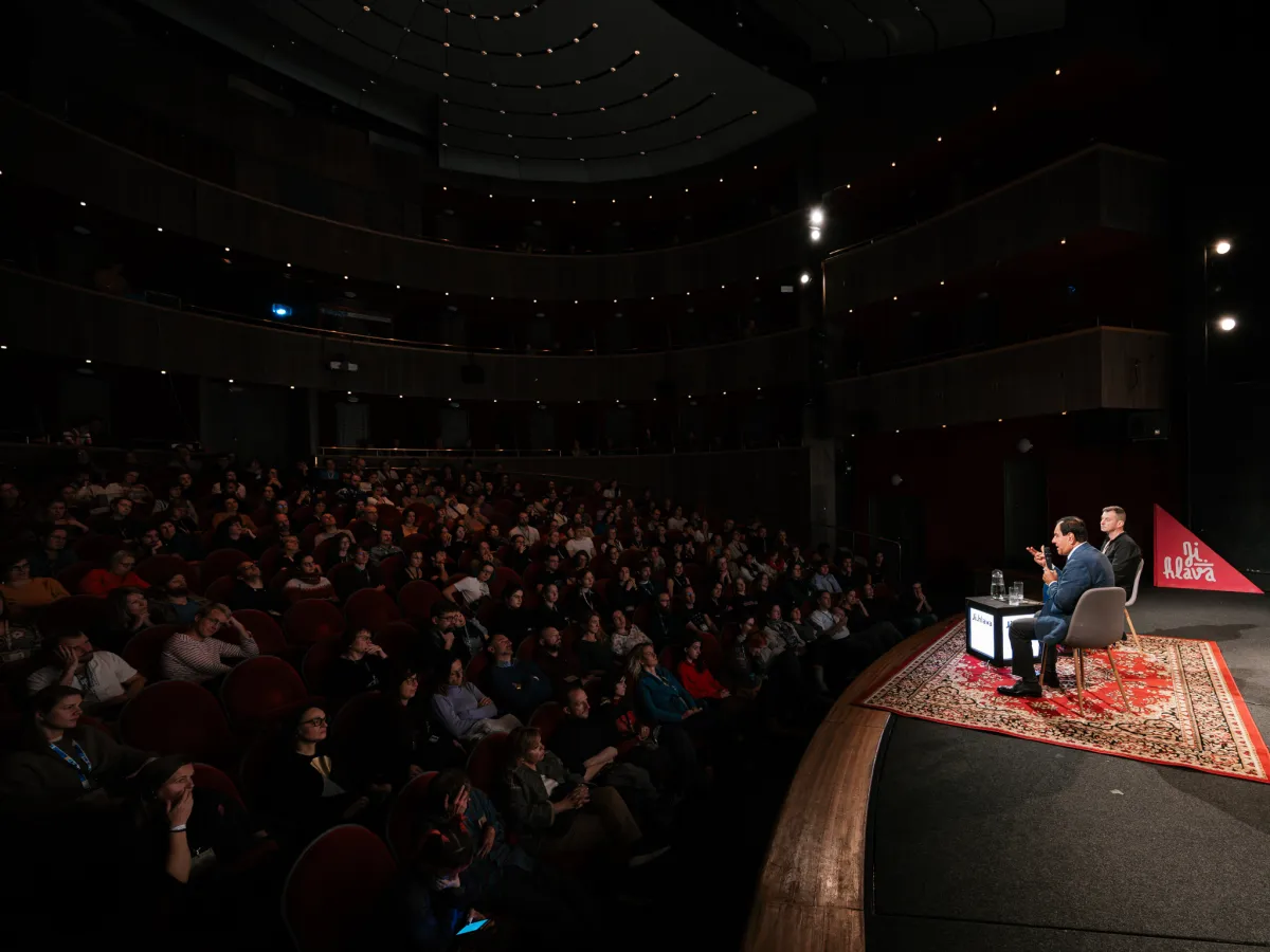 At a stage, two men sit in chairs as a rapt theater audience looks on