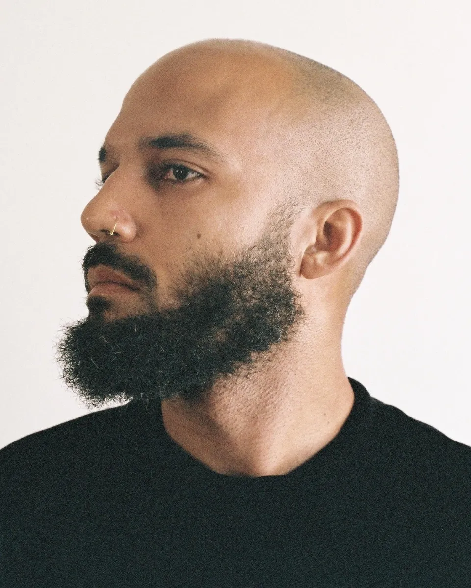 A headshot of a light-skinned Black bald man with full black beard and a thin nose ring wearing a black t-shirt, seen in profile,