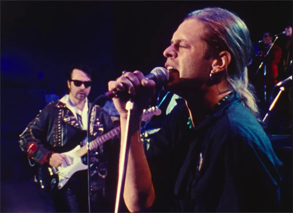 A man with a blond ponytail sings passionately into a microphone, in front of the rest of his rock band.