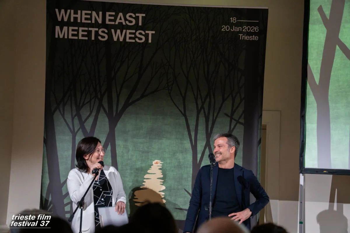 A light-skinned woman with dark hair and glasses, wearing a black shirt and a white cardigan talks into a microphone while talking to a white man in a black sweater with short-cropped hair with a poster advertising When East Meets West