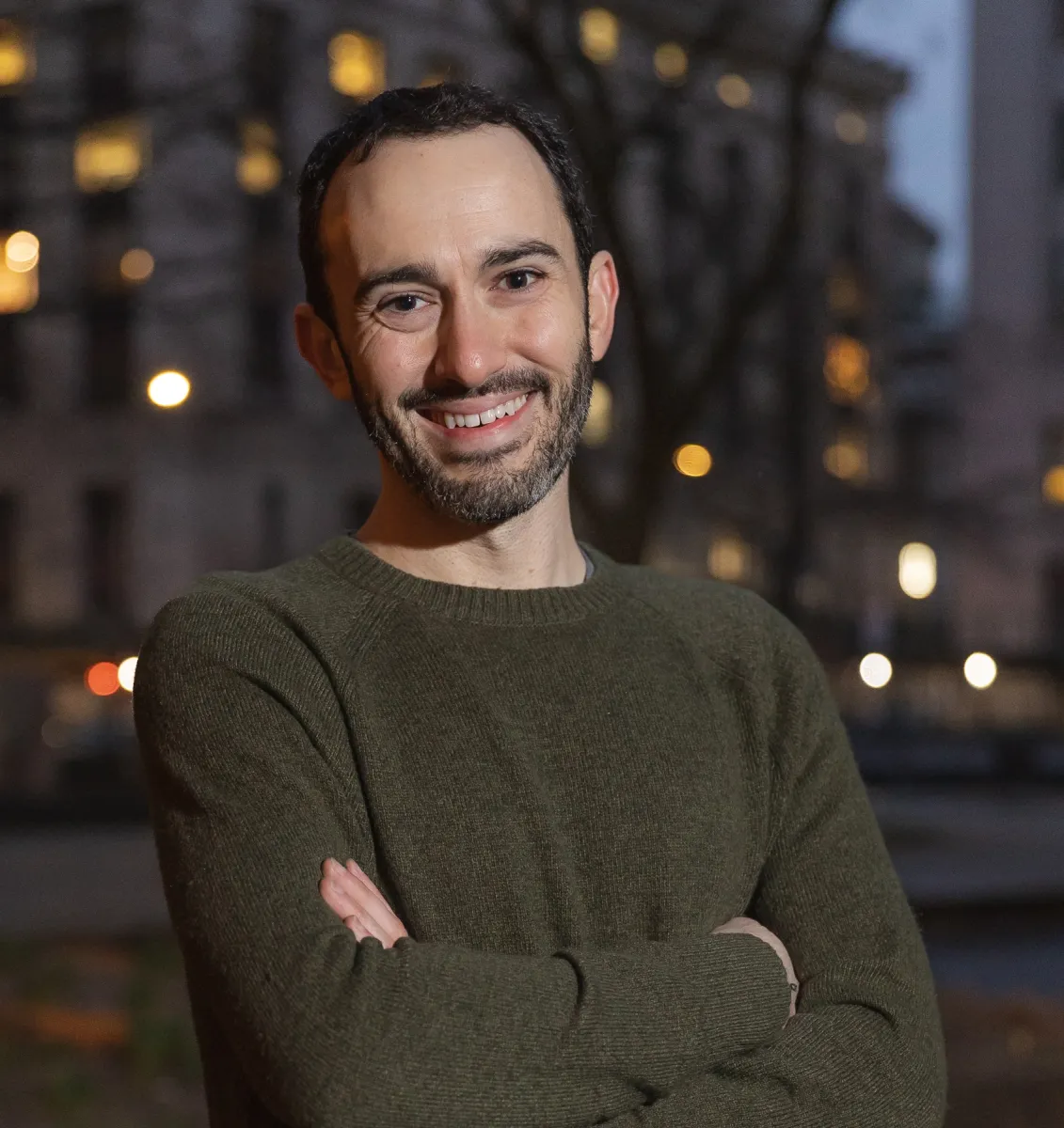 Headshot of a smiling young bearded white man with short dark hair in a city street