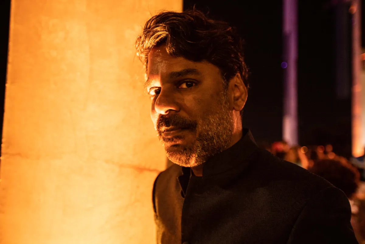 A headshot of a dark-skinned man with thick head of hair and stubbled beared wearing a black shirt, lit at night by an orange glow