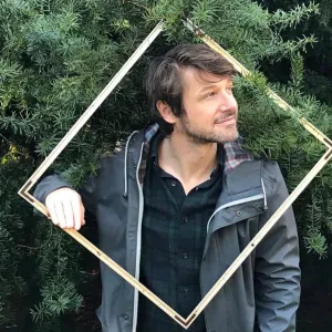 A young white man with flowing brown hair and stubble wears a black shirt and a jacket outside with a frame