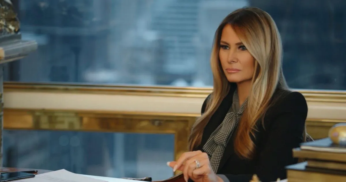 A heavily-made up white woman with long, colored blond hair sits a desk with a stern look on her face