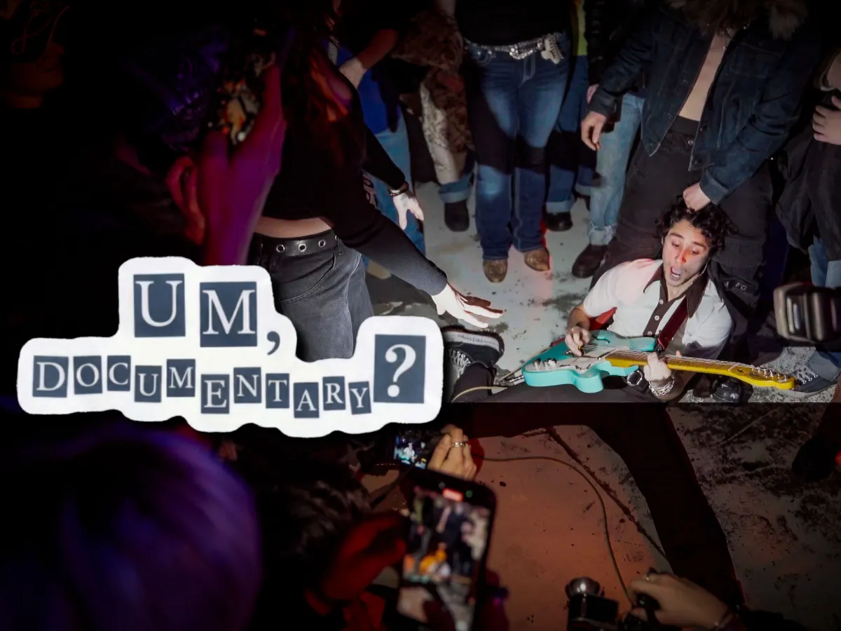 Eli, the frontman of Um, Jennifer?, is on the ground playing a blue guitar. He is surrounded by a swarm of energetic fans recording and cheering him on. The image is captured from an Um, Jennifer? show in Brooklyn. He is wearing gray trousers and a white blouse.