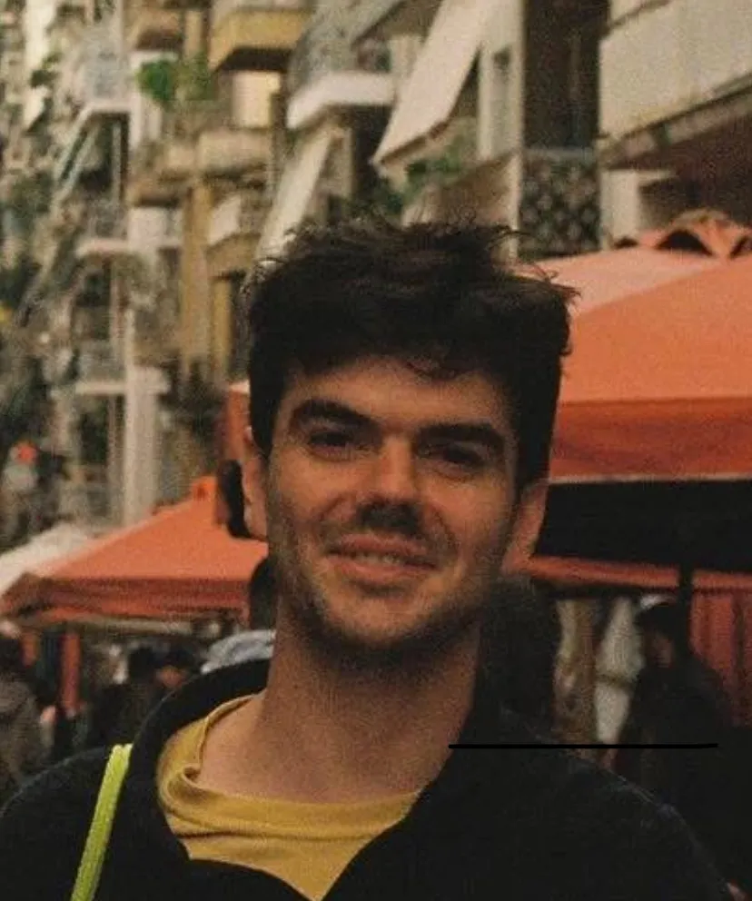 Blurry, grainy headshot of a young white man with short dark hair and 5 o'clock shadow wearing a yellow t-shirt and a black sweater