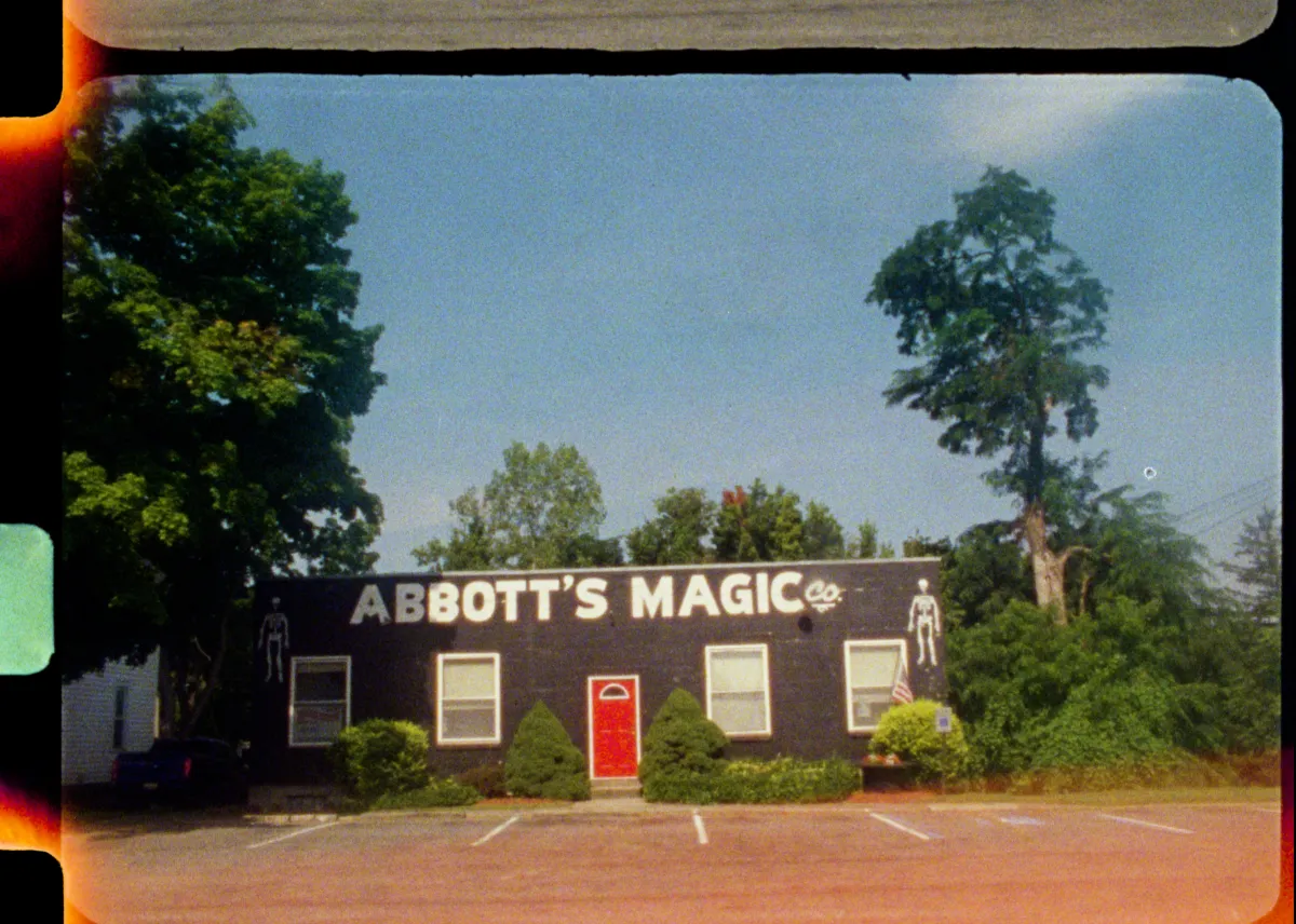 a 16mm film still of a low, black painted brick building with skeletons painted on the front labeled "Abbot's Magic" in large white lettering