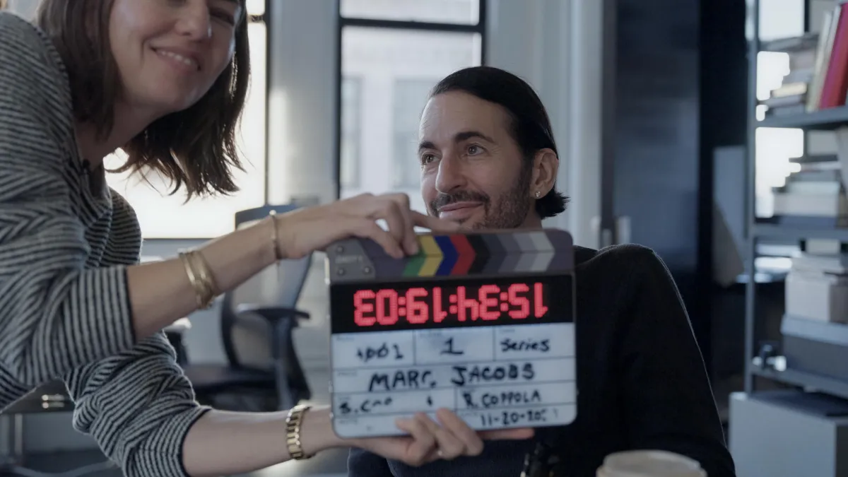 A middle-aged white man with slicked back black hair and five o'clock shadow sits in front of a camera as a white woman with shoulder-length brown hair holds a clacker that reads "Marc Jacobs" in front of him
