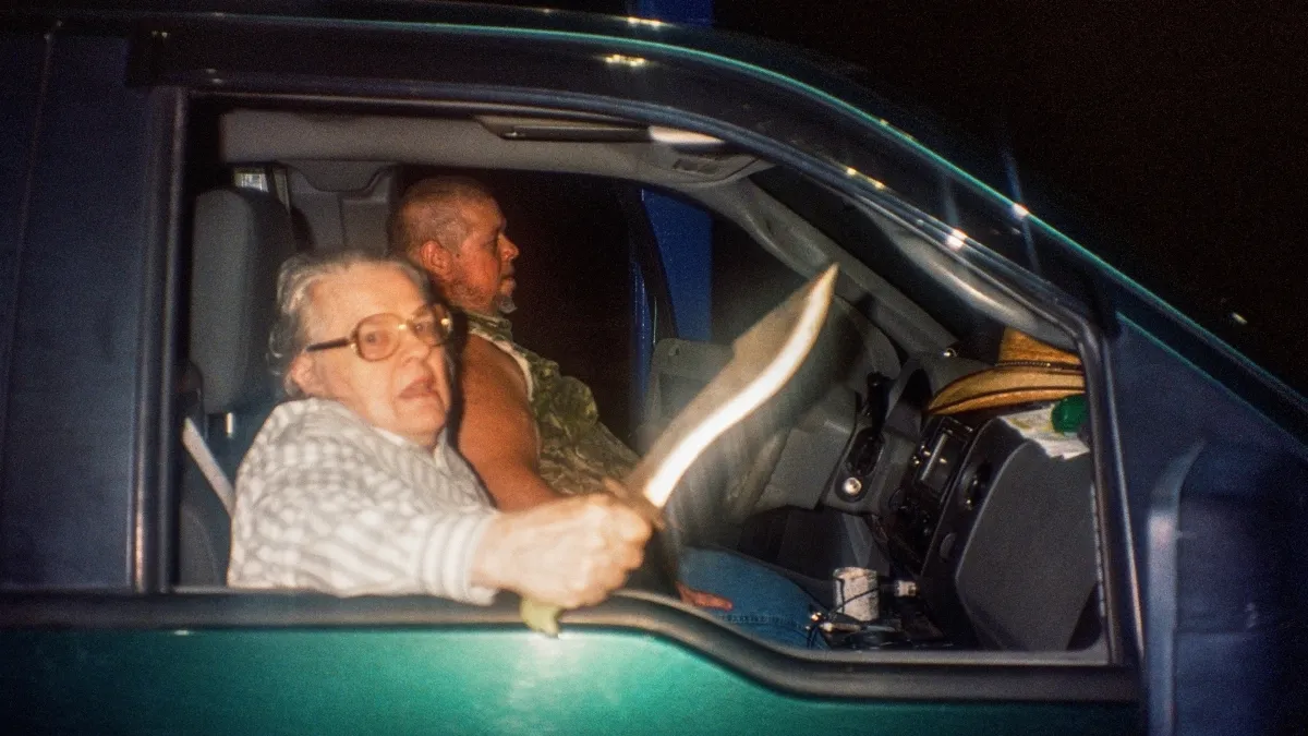 An older women with glasses sitting in a truck at night wields a knife at the camera
