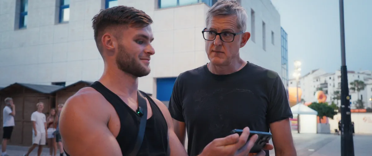 A middle-aged white man in a black t-shirt and rimmed glasses stares incredulously at a young muscled white man in a black tank top as he stares into his smartphone