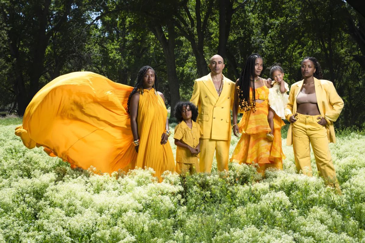 A family of six stands together in a sunlit field of soft white wildflowers, surrounded by lush green grass and tall, dark trees. At the center, a man and woman stand close, with four children gathered around them—one held in the woman’s arms, others standing confidently at their sides. All are dressed in coordinated, flowing and tailored outfits in vibrant shades of yellow and gold, creating a striking contrast against the deep green landscape. One figure on the left wears a dramatic, billowing gown that e