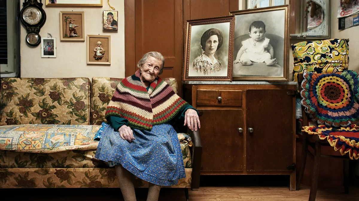 An elderly woman with pulled back grey hair sits on a yellow-print heavy coach surrounded by framed black and white photos of her younger self 