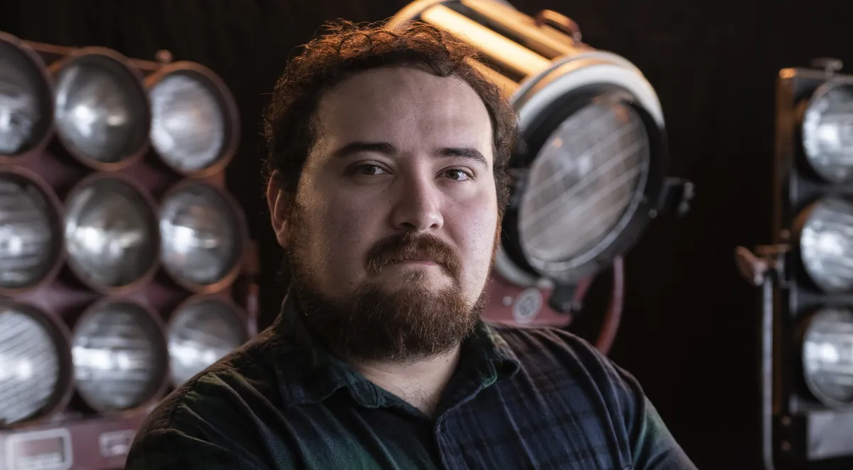 A young light-skinned man with short brown hair and a red-tinged brown beard stares at the camera standing in front of movie lights