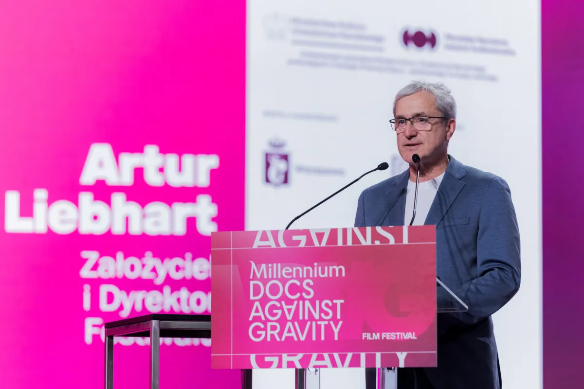 A middle-aged white man with grey hair and glasses wears a suit and a tee while standing at a podium that's branded as Millennium Docs Against Gravity in bright pink