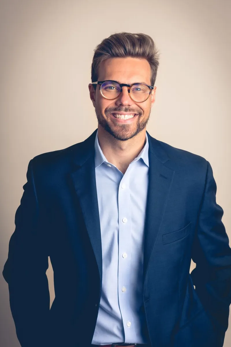 Headshot of a white man with well-coiffed light brown hair wearing a light blue shirt, dark blue blazer, donning glasses and a big smile