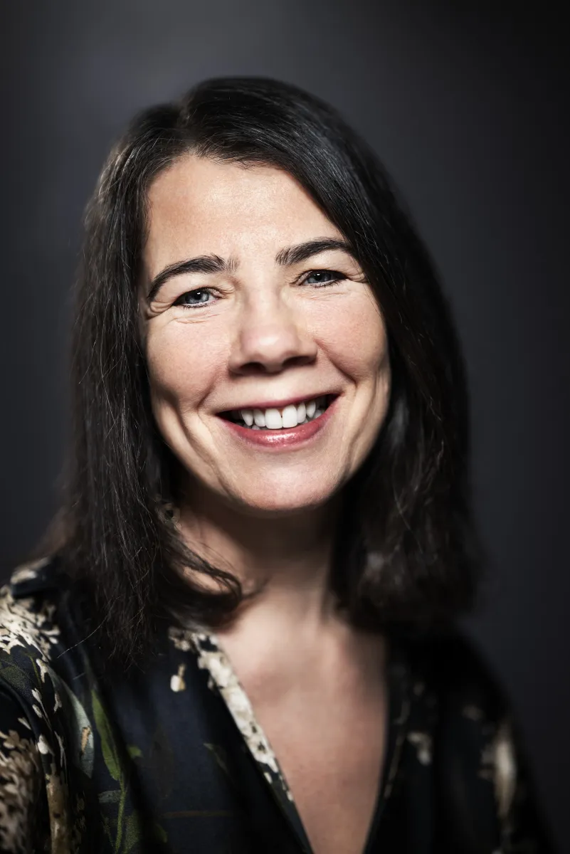 Headshot of a smiling white woman with straightened shoulder-length black hair and a v-neck black blouse