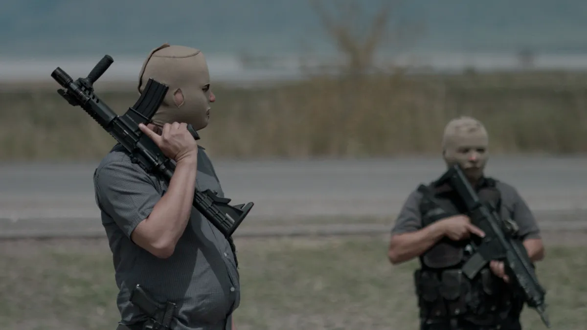 Two men in cammo gear stand in an empty field holding guns don skin-colored masks that reveal only their eyes, nose, and mouth