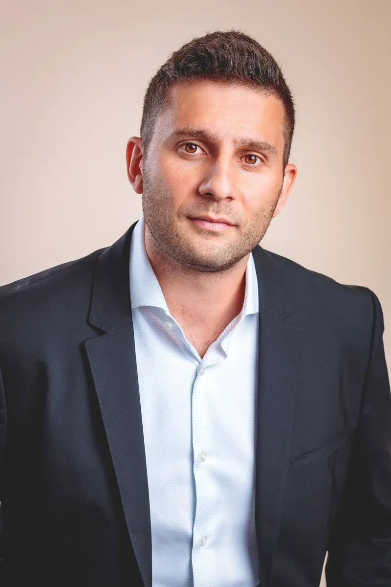 Headshot of a light-skinned man with short-cropped dark brown hair wearing a light blue shirt and dark blazer