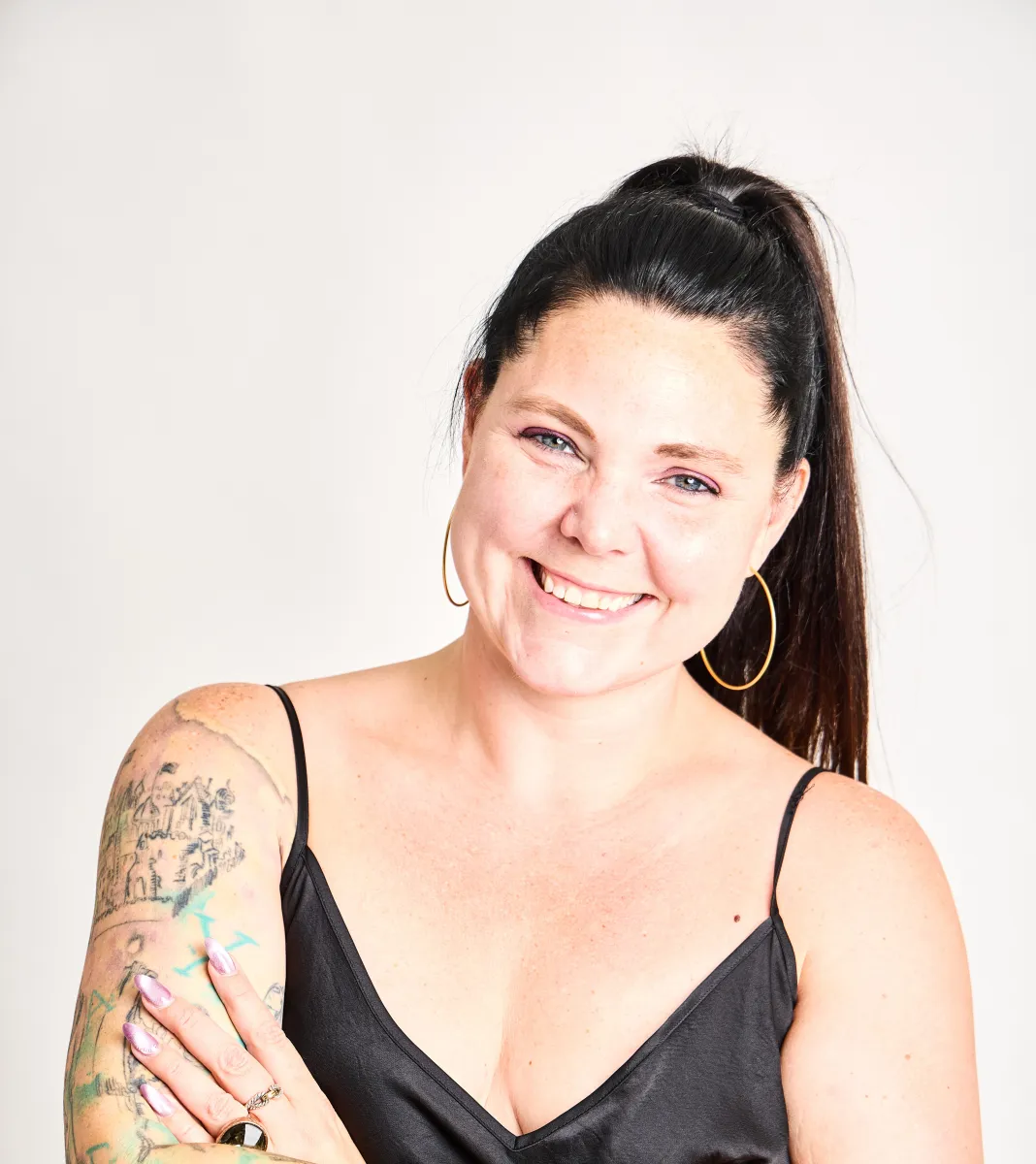 Headshot of a white woman wearing a black top with spaghetti straps that show off her arm tattoos, her black hair up in a ponytail, smiling