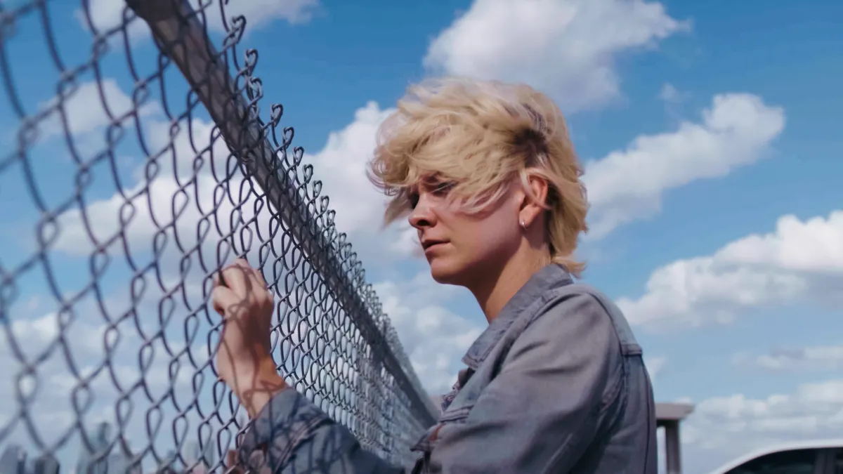  a young person with short, windblown blond hair standing beside a chain-link fence under a bright blue sky with large white clouds. They are wearing a gray denim-style jacket and appear to be looking through or past the fence with a quiet, reflective expression.