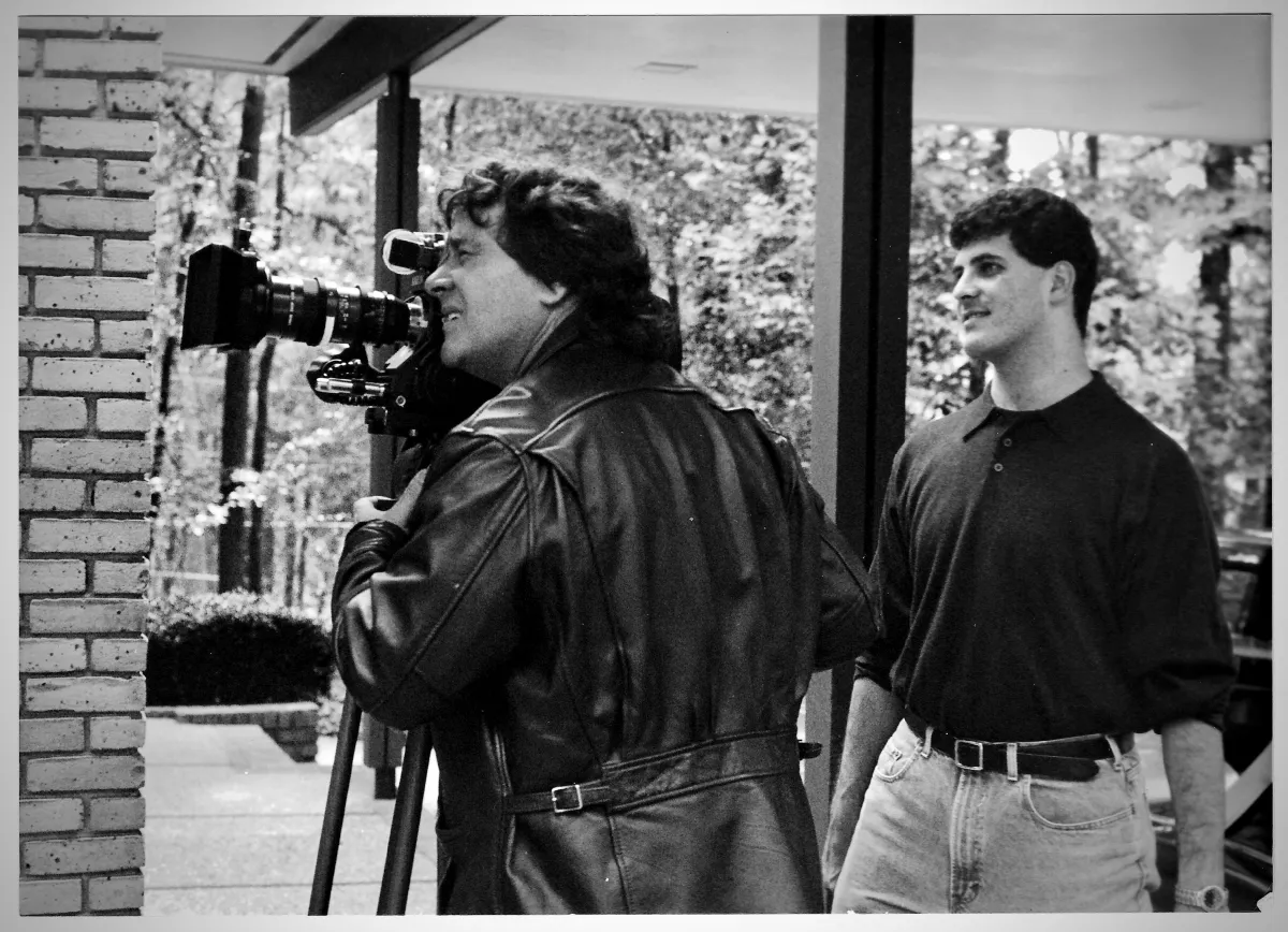 On the left, a man in a black leather jacket peers into a video camera. On the right, a younger man in a tucked polo observes.  Black and white photo