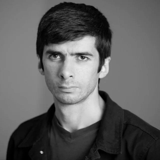 Black and white headshot of a light-skinned man with short dark hair wearing a dark collared shirt over a dark tee furrowing his brow at the camera