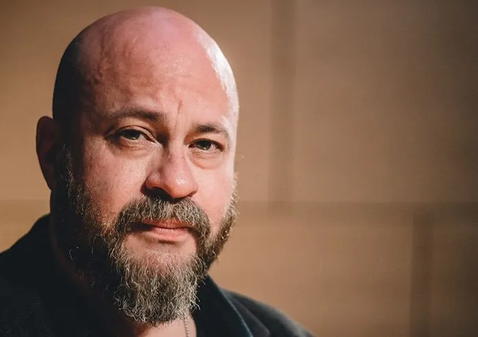 Headshot of a light-skinned bald man with a bushy beard furrowing his brow at the camera
