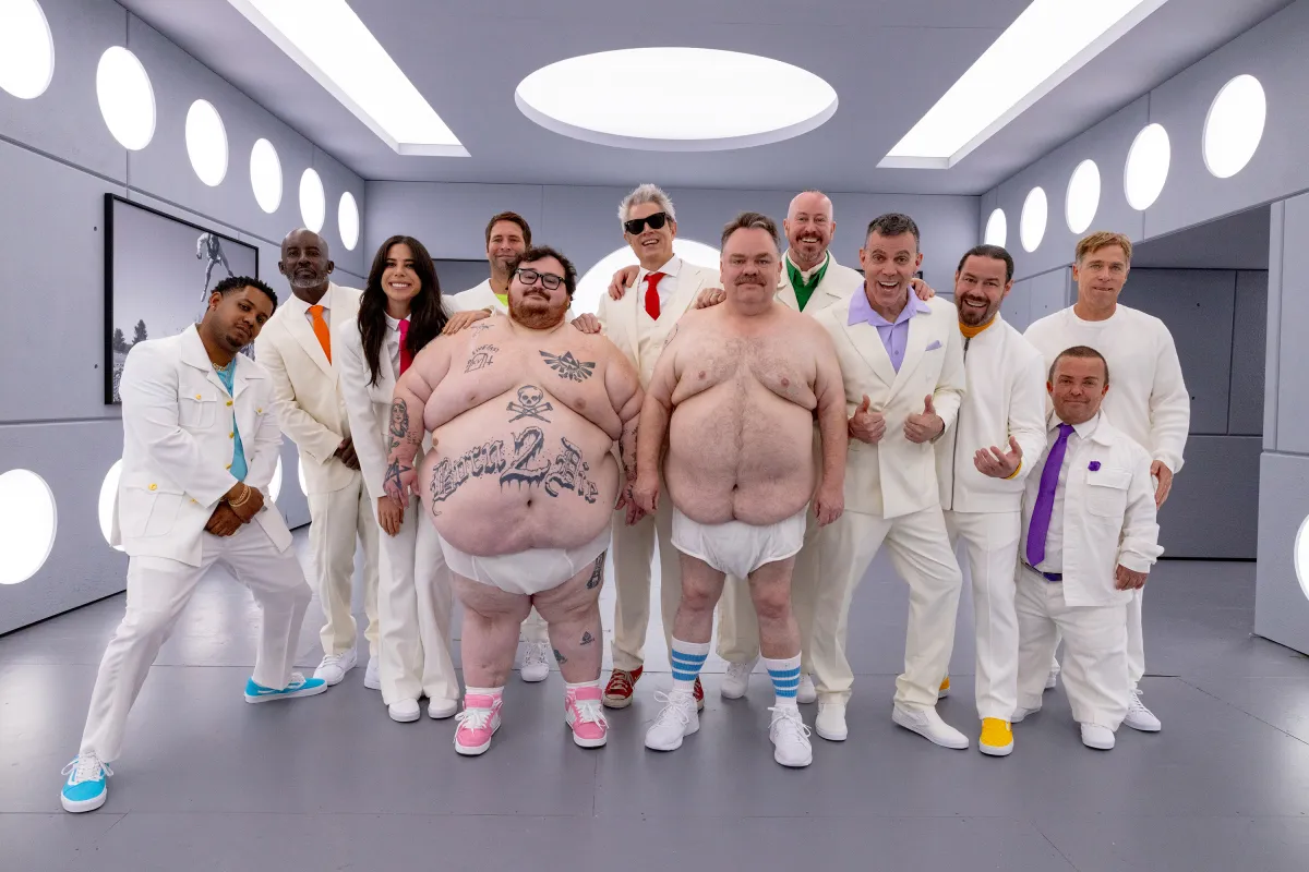 Two large men wear nothing but sneakers and diapers. One is decorated in tattoos, including a skull and crossbones and text which reads "BORN 2 DIE." Nine men and one woman stand behind the two in matching white suits. The ensemble appears in a sterile, grey room with futuristic, circular white lights