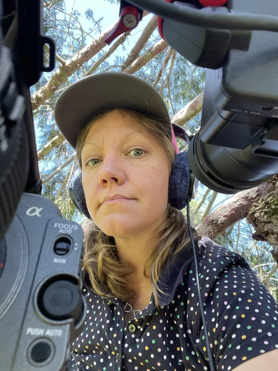 Young white woman with blond hair in a polka dot shirt and cap, wears headphones amid cameras looking down at us