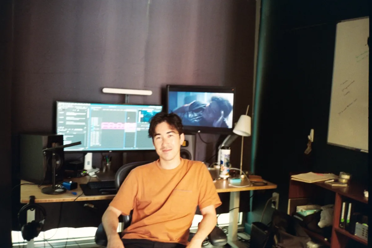 Young man in orange shirt sits in front of editing workstation with two large monitors