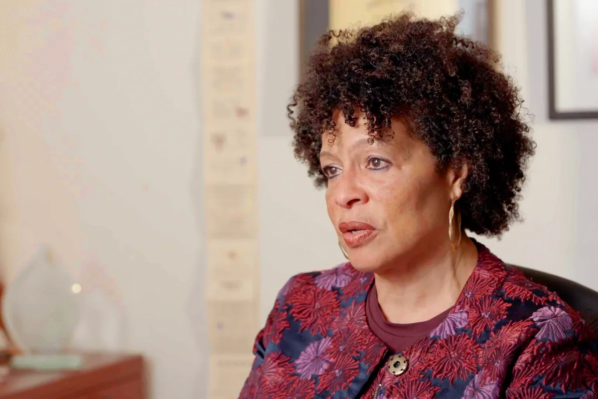 African American woman in her fifties, seated at a desk wearing a top featuring red and pink flower design. Picture frames are visible, but out of focus, in the background.