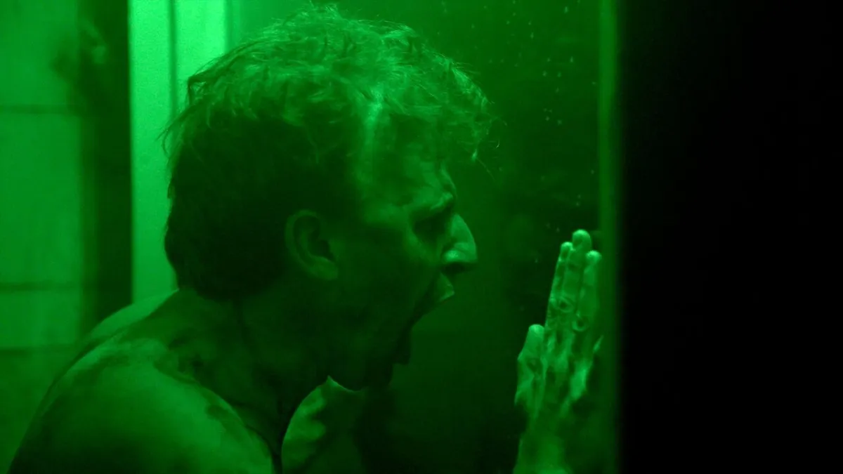 A white man presses against glass and screams, lit in bright green