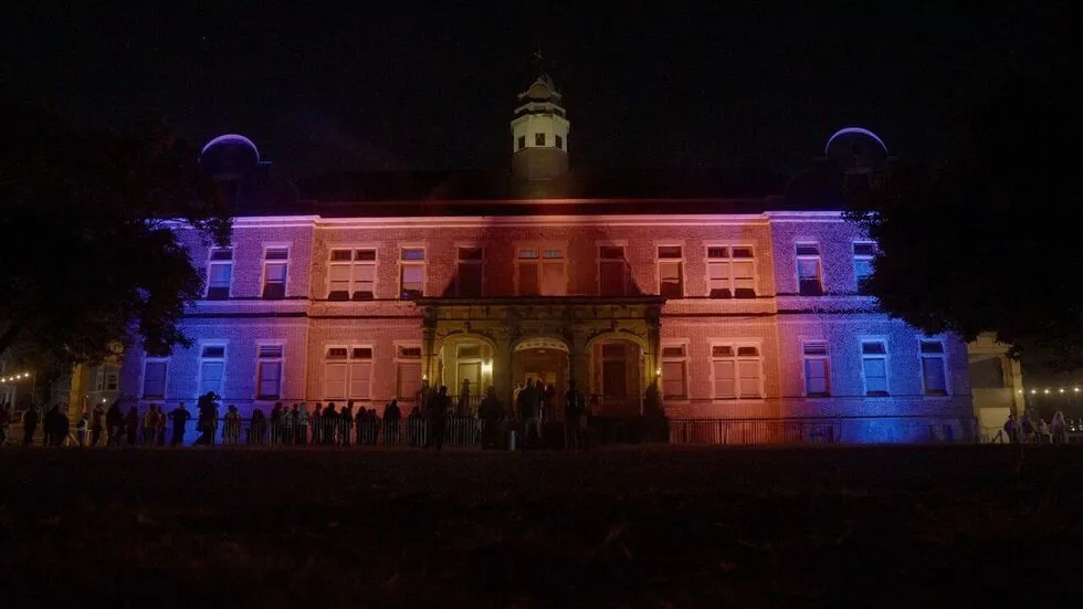 A long line of silhouetted figures stand in front of a large university building lit in orange and blue at night