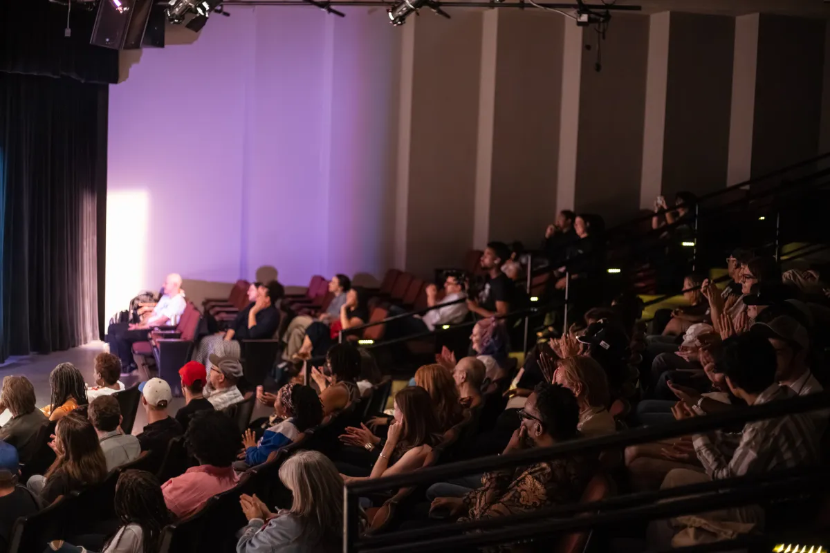 A packed theatre focuses on a stage which is off camera to the left. Purple light illuminates the audience