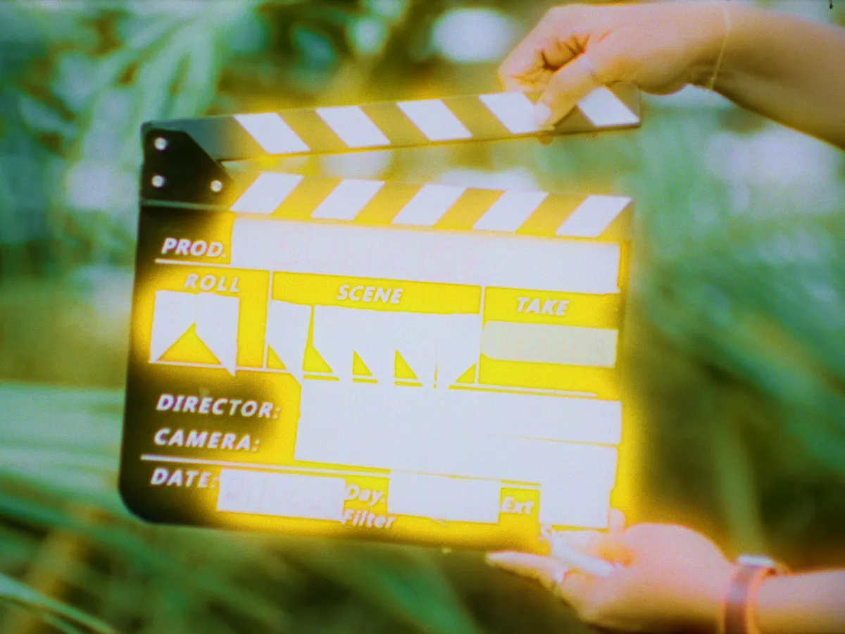 Two hands prepare to clap shut a slate in front of ferns. The text on the slate is obscured by shining yellow light