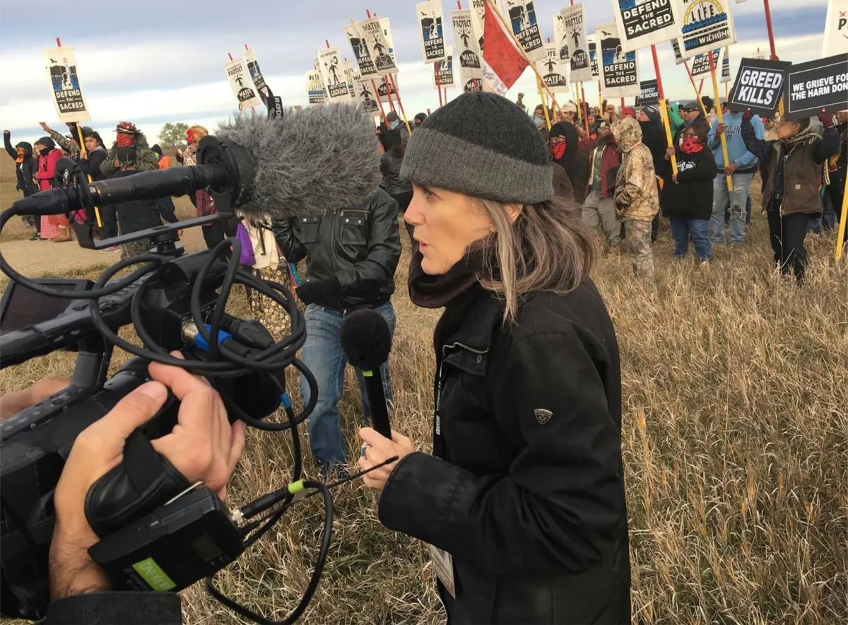 A middle-aged white woman in a black jacket and a grey wool hat stands in front of a protest holding a mic speaking into a camera