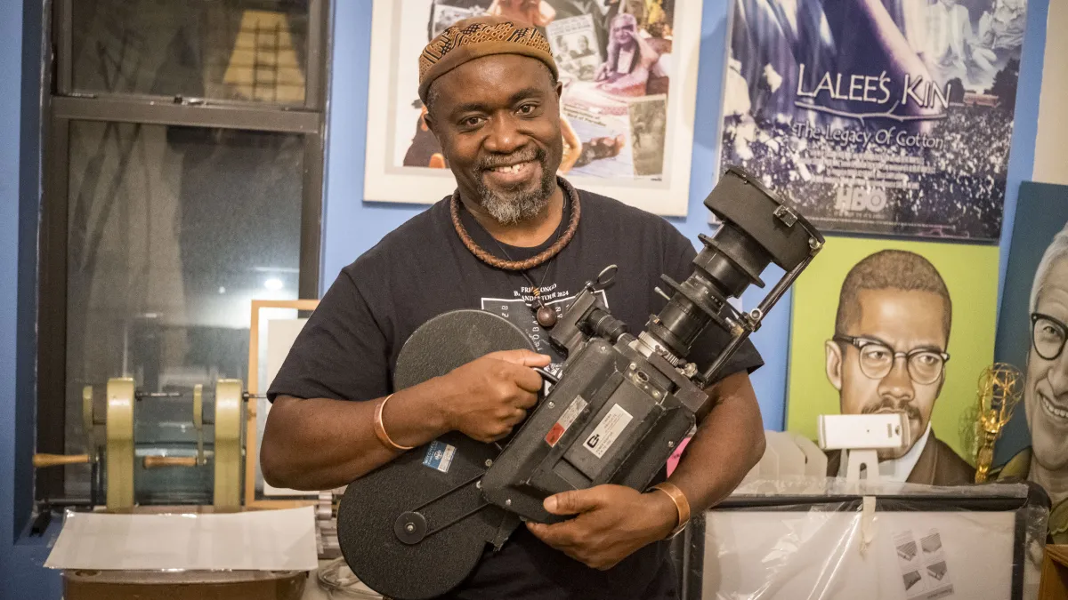A smiling Black man in a brown hat, black T-shirt, and wooden and metal jewelry holds an old camera in front of awards and posters, one depicting Malcolm X