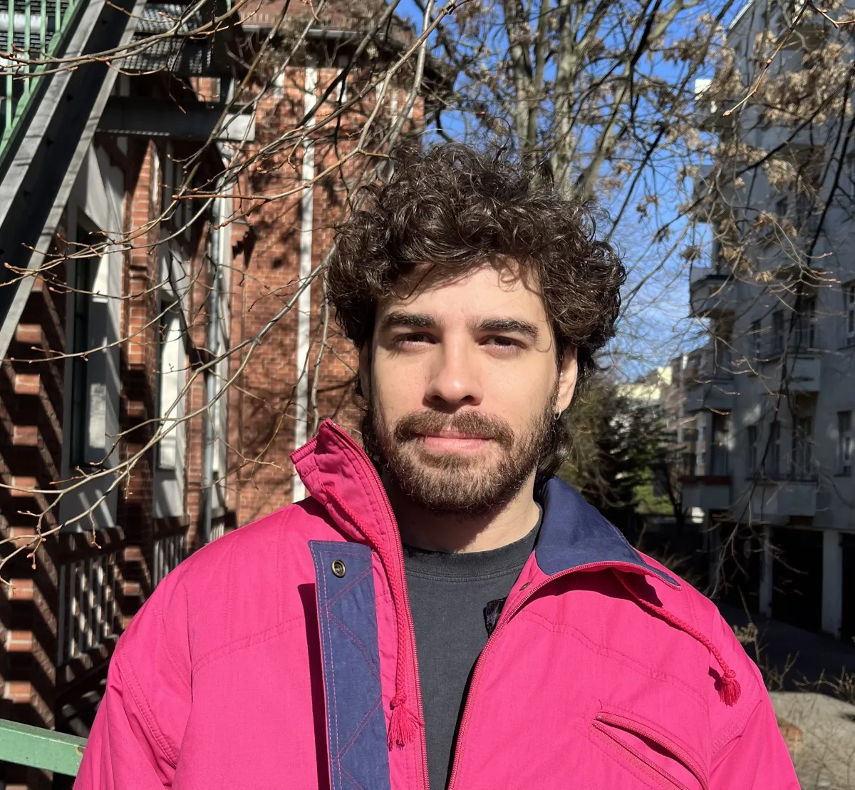 Headshot of a man with brown facial hair and curly hair wearing a pink jacket over a black T-shirt. A city is in the background