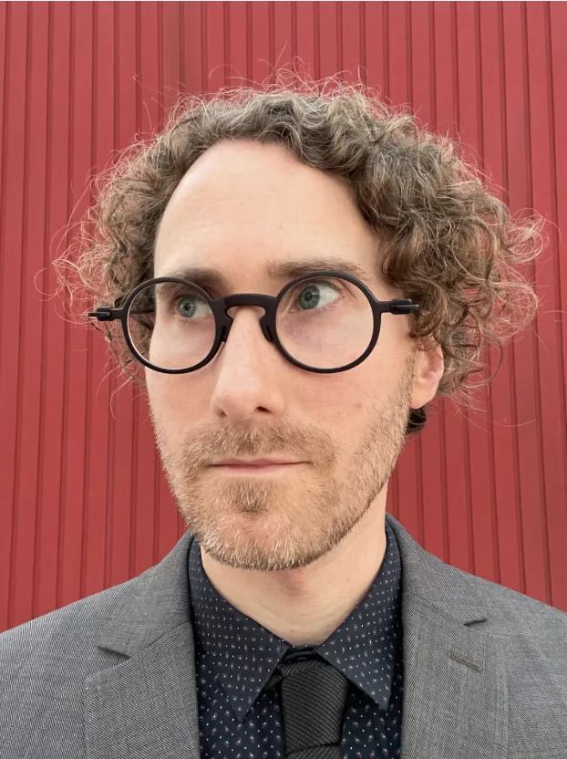 A light-skinned man wearing dark glasses is looking to the left with a timid smile. He has dark blond, curly hair. He is dressed in a gray suit and a black tie, with a red background behind him.