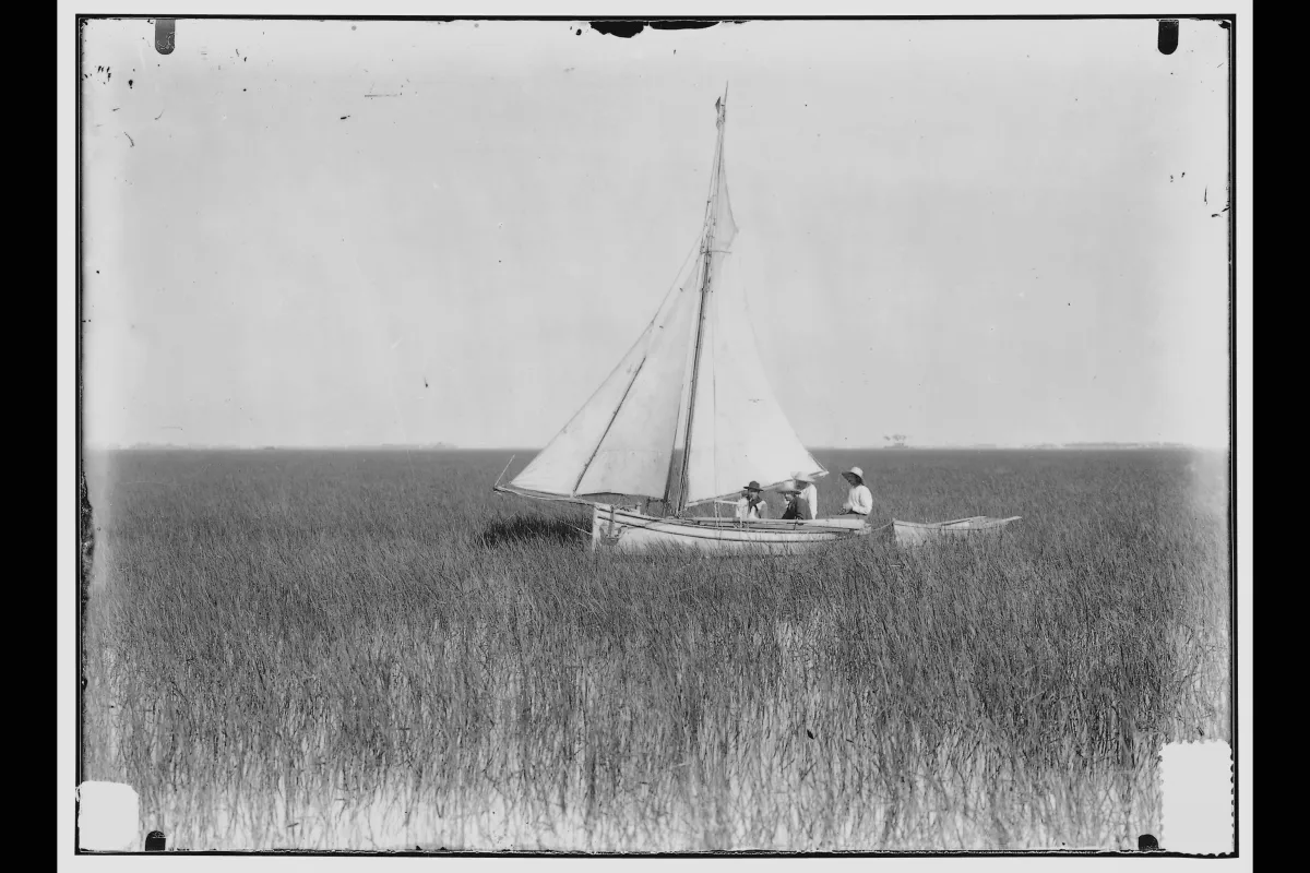 Black and white image of three men sitting in a white sailboat amidst marshland