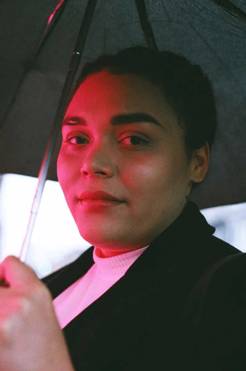 Headshot of a Black woman in a black jacket and white shirt holding a black umbrella, illuminated by red light