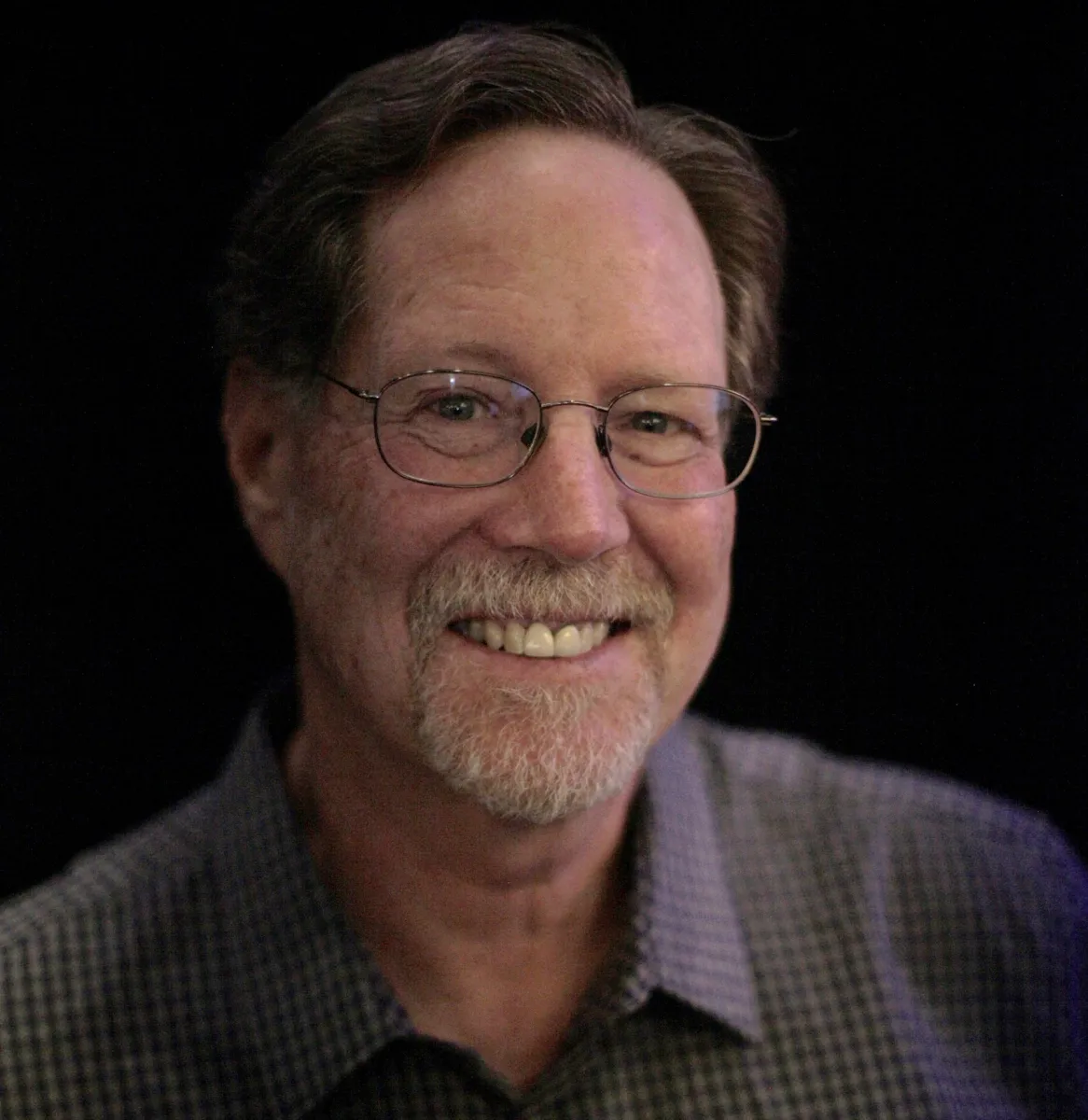 Headshot against a black backdrop of middle-aged white man with glasses, brown hair and short white beard wearing a purple-hued shirt.