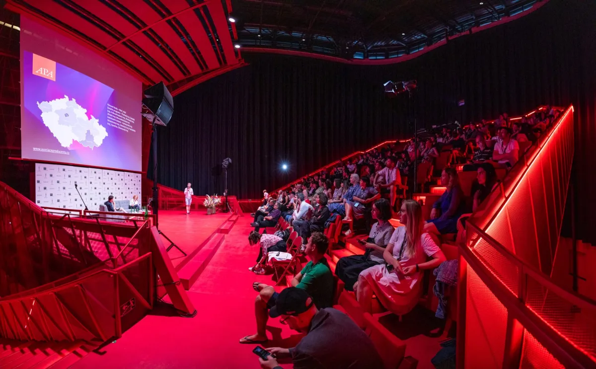 A large crowd in a theater lit in red looks at a big screen which displays a map of Czechia over a purple background