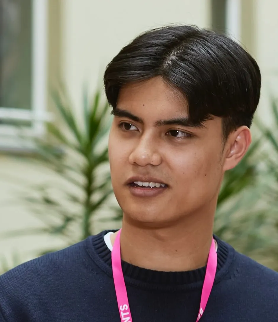A young dark-skinned man with black parted hair wears a blue sweater and a pink lanyard around his neck