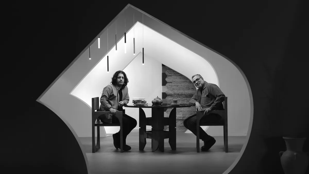 Two men sit at a table in a modern room framed by an unusually shaped doorway. Both look towards the camera. The man on the left has shoulder length dark hair and the man on the right has short greying hair and facial hair and glasses. Two mugs of coffee, a stack of books, and three decorative vases sit on the table. An abstract chandelier appears above the men's heads. Black and white image
