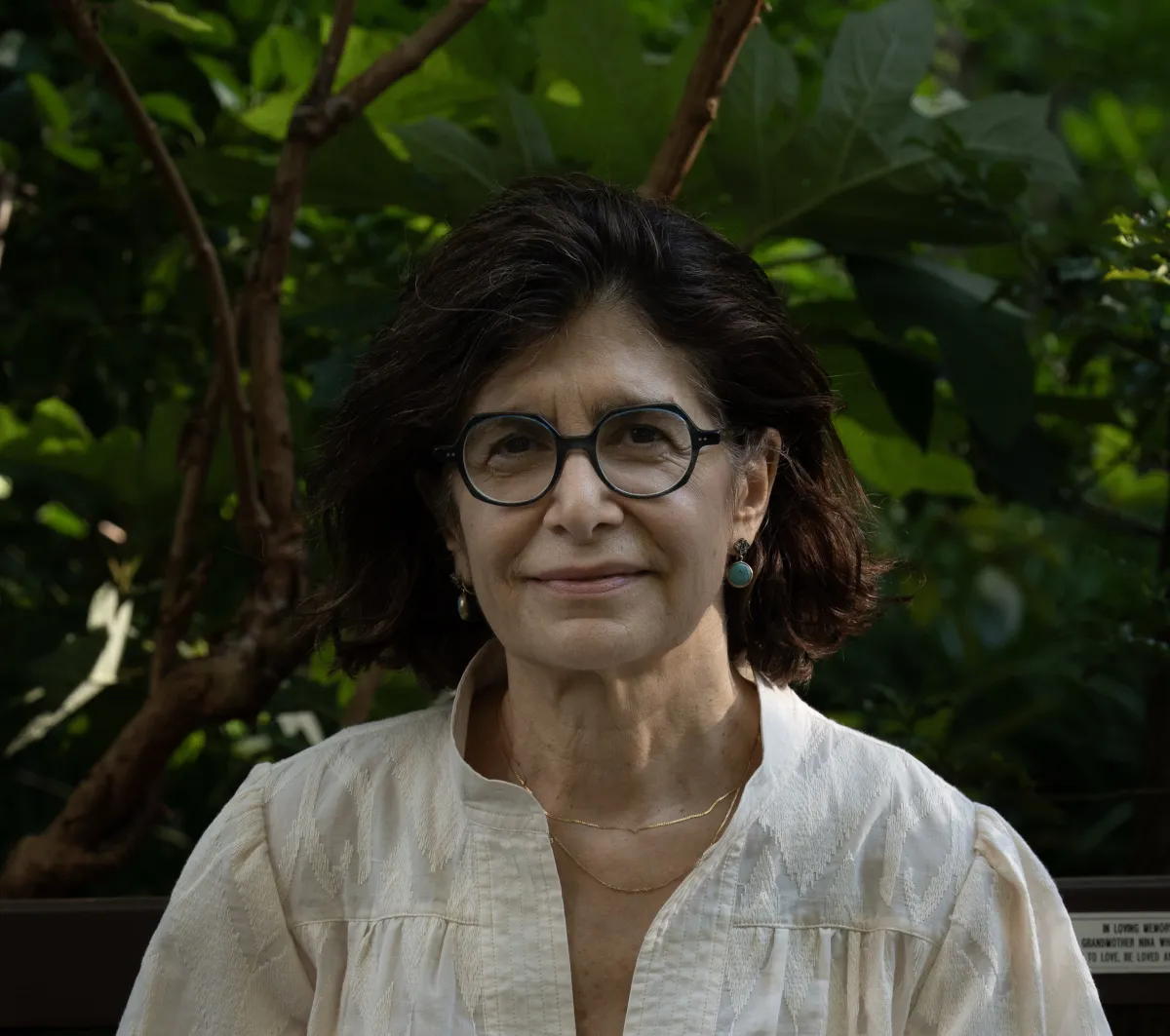 Dimly lit headshot of a smiling woman with shoulder length dark hair in a white blouse and large glasses in front of a tree