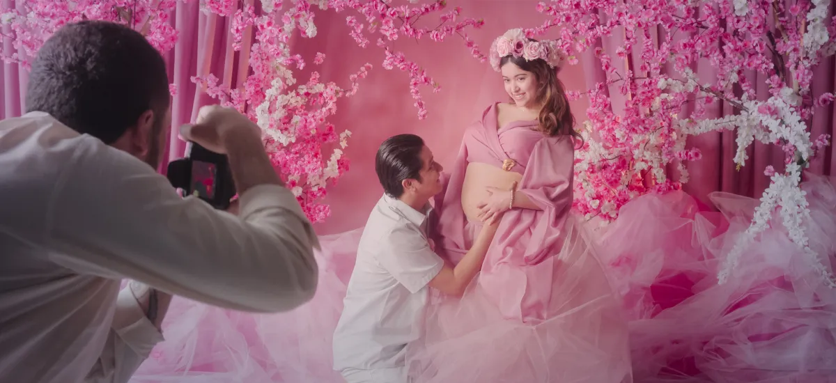 A photographer in frame right photographs a pregnant woman in a pink dress and pink flower crown in front of a pink background. A man in white kneels and kisses her stomach