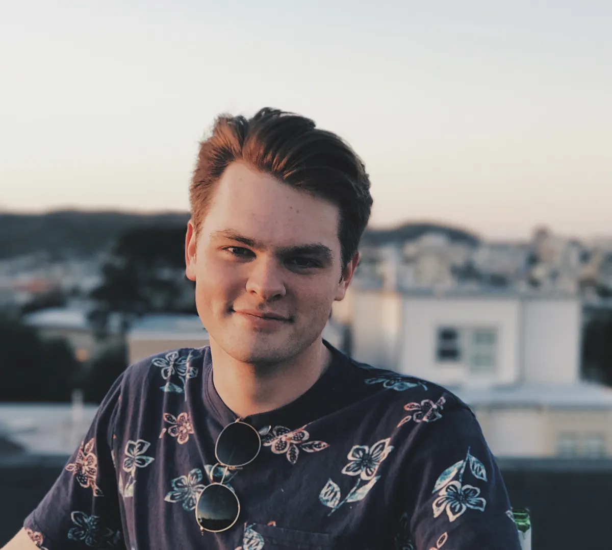 Headshot of a white man with short, combed-back brown hair smiling in front of blurred white buildings. The sun sets on his face and he wears a navy T-shirt with a floral pattern